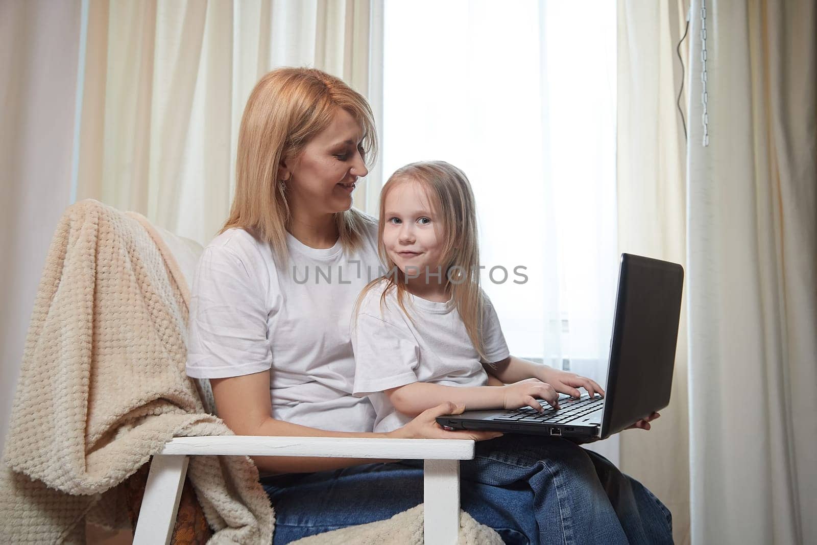 Beautiful young woman and her little cute daughter are using laptop at home. Enjoying spending time together with Internet and modern technologies