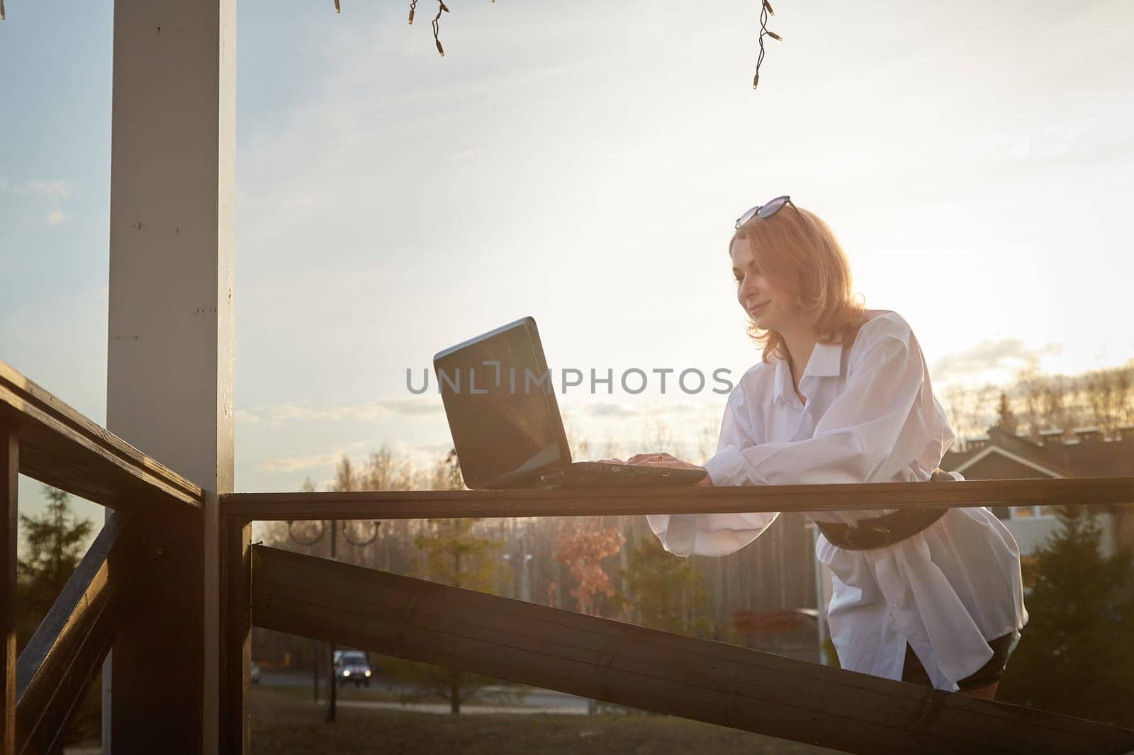 Beautiful redhead moden girl working by laptop in gazebo on autumn, summer or spring day. Businesswoman, student, freelancer or manager works outdoors in natural landscape in village, city or town by keleny