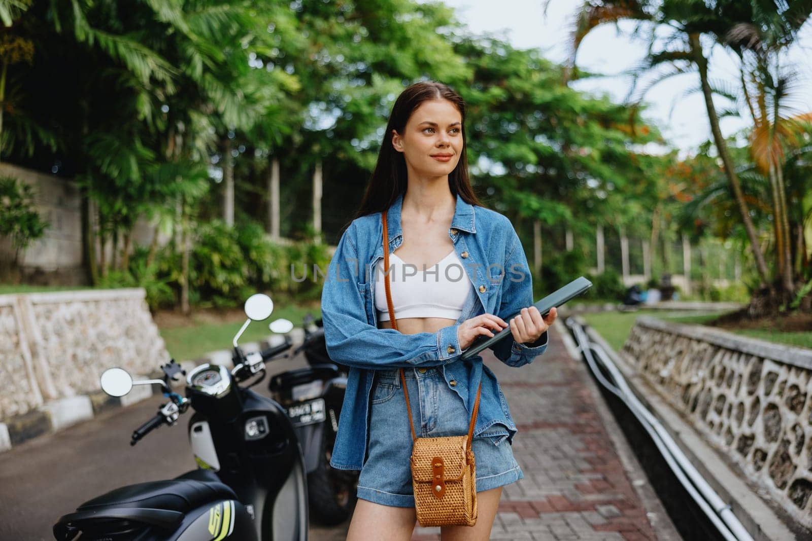 Woman smiling walking in the park outside with laptop freelancer against a backdrop of green palm trees in summer, tropical backdrop, blogger on a trip, work online by SHOTPRIME