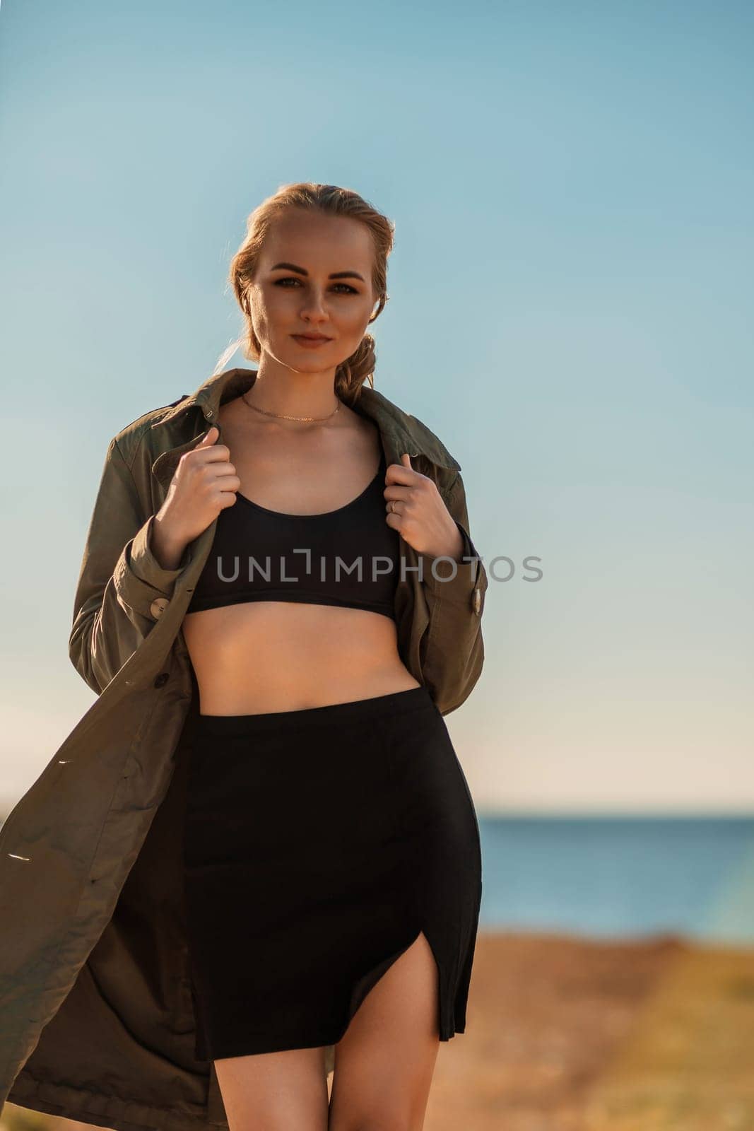 Portrait blonde sea cape. A calm young blonde in an unbuttoned khaki raincoat walks along the seashore, under a raincoat a black skirt and top.