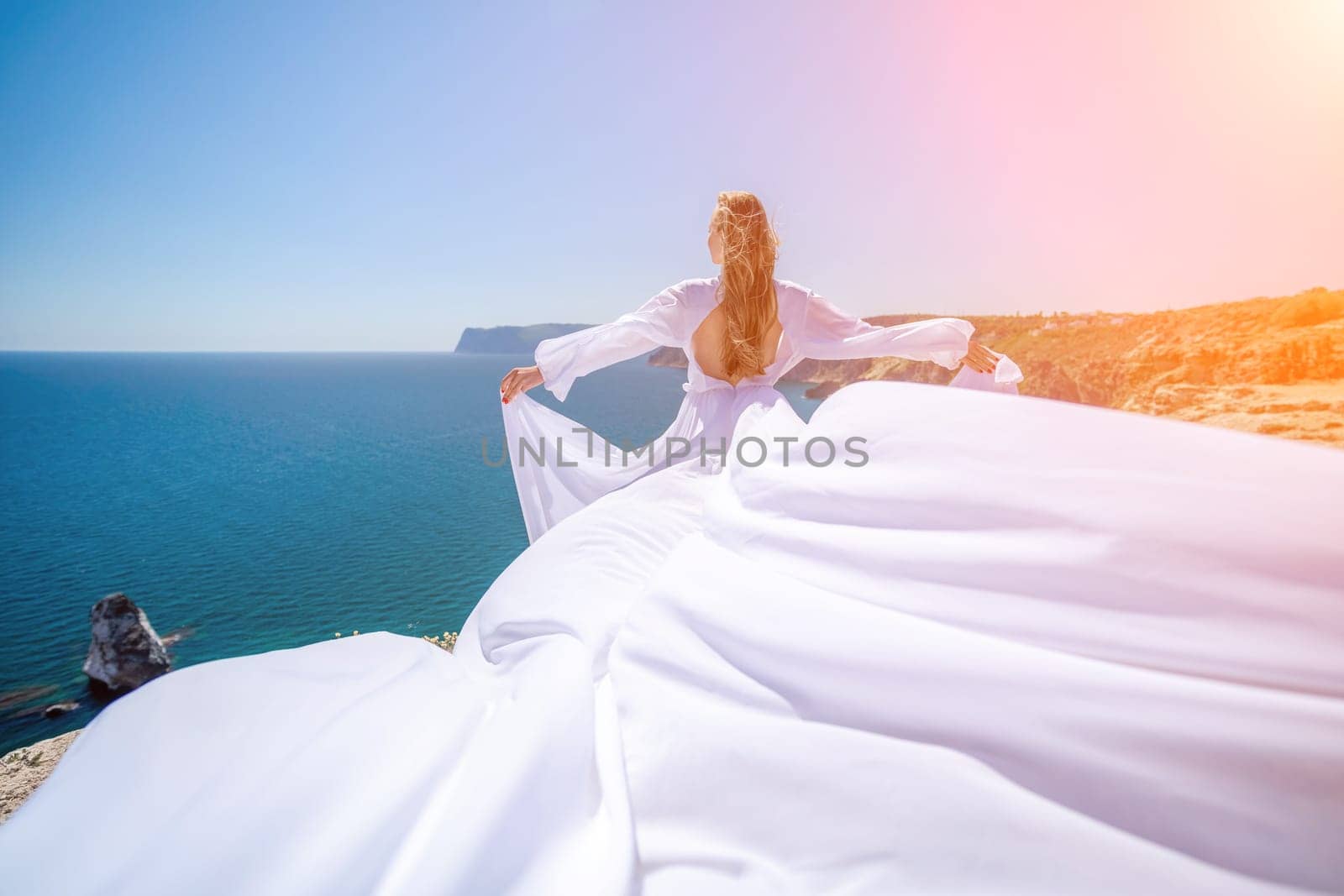 woman sea white dress. Blonde with long hair on a sunny seashore in a white flowing dress, rear view, silk fabric waving in the wind. Against the backdrop of the blue sky and mountains on the seashore