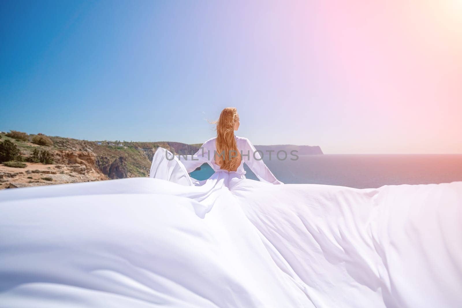 woman sea white dress. Blonde with long hair on a sunny seashore in a white flowing dress, rear view, silk fabric waving in the wind. Against the backdrop of the blue sky and mountains on the seashore