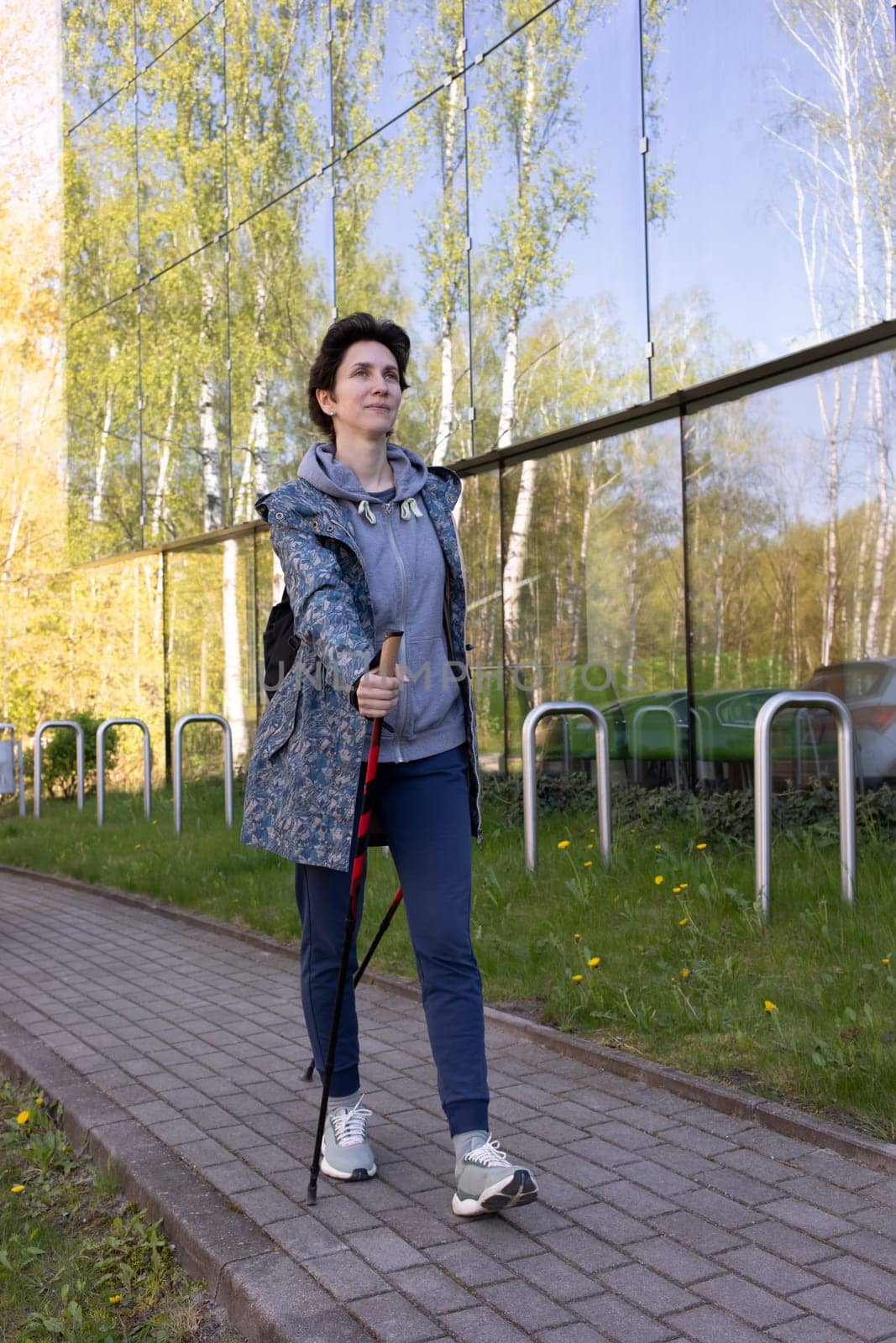 Nordic Walking In Urban City. Beautiful Happy Caucasian Woman Training With Sticks Outdoor, Modern Glass Eco Building on Background. Physical Activity With Walking Poles, Recreation. Vertical Plane.