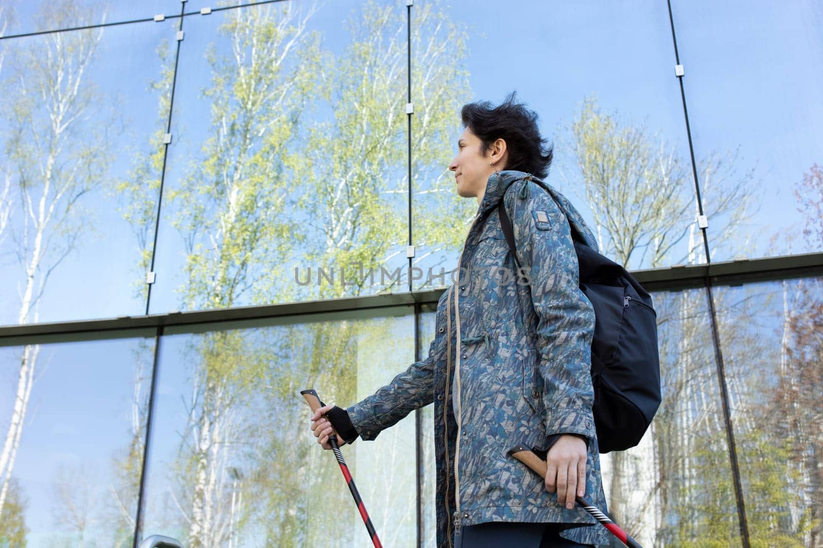 Pole Walking In Urban City. Beautiful Happy Caucasian Woman Training With Sticks Outdoor, Modern Glass Eco Building on Background. Physical Activity, Nordic Walk, Recreation. Horizontal Plane.