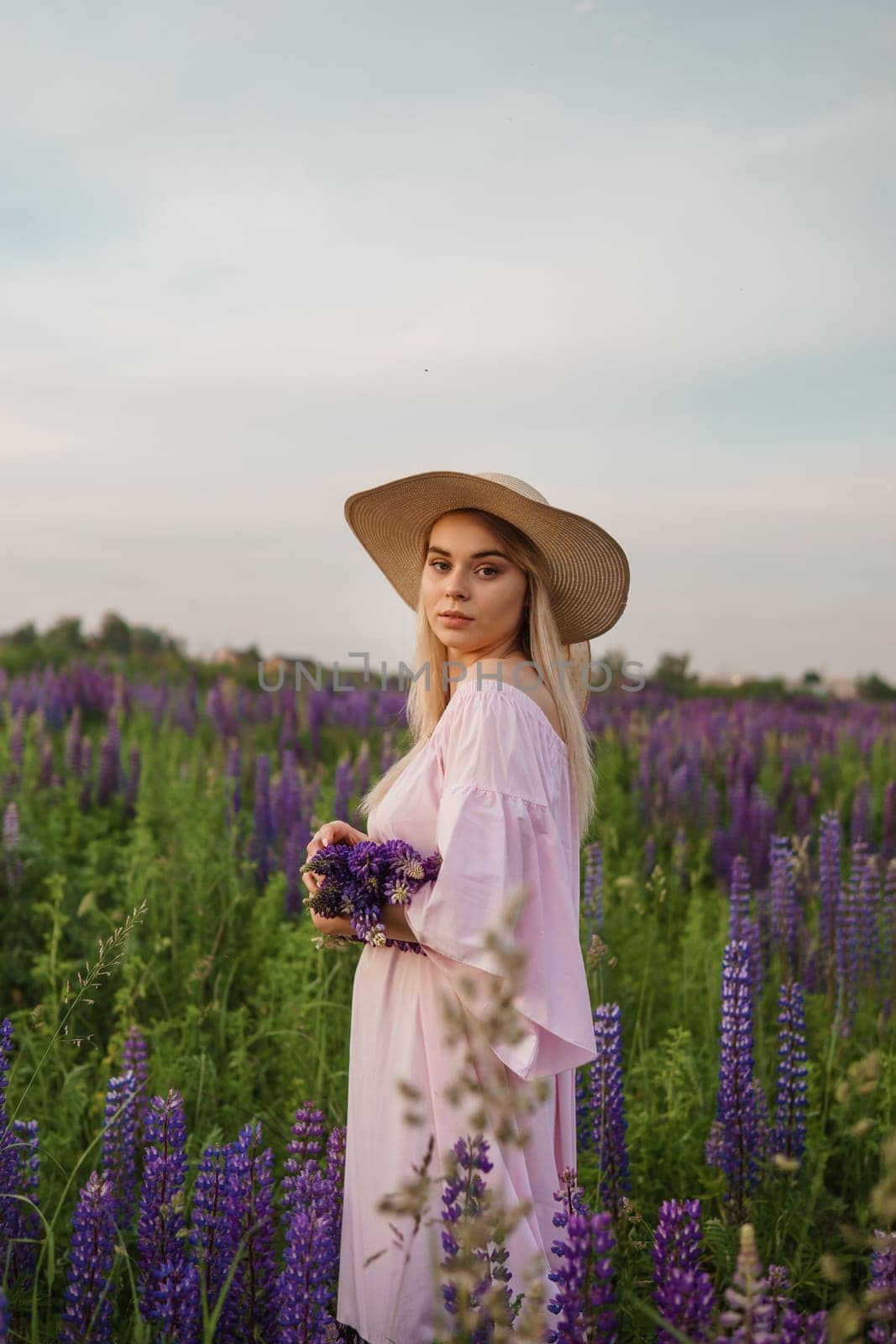 A beautiful woman in a straw hat walks in a field with purple flowers. A walk in nature in the lupin field.