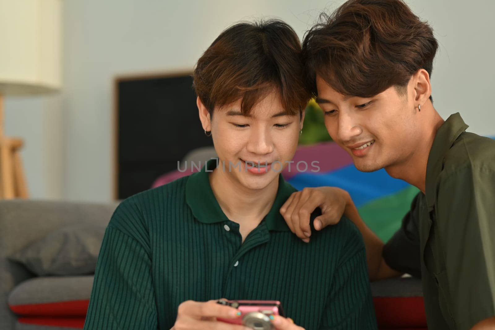 Joyful gay couple taking a selfie with compact camera in living room rainbow flag in background. LGBT, love and human rights concept by prathanchorruangsak