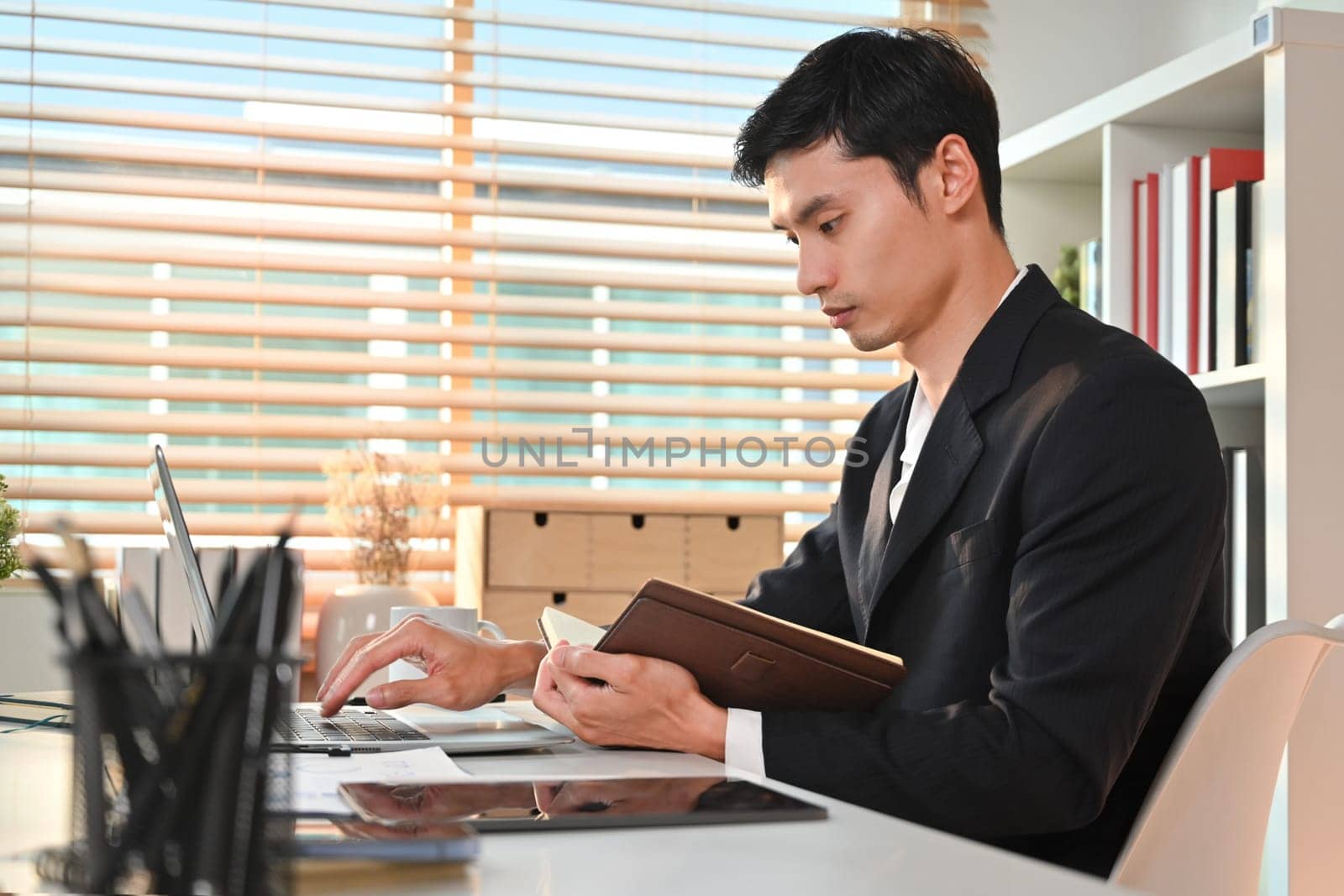Side view of millennial male entrepreneur using laptop computer and checking schedule on his diary at his office by prathanchorruangsak