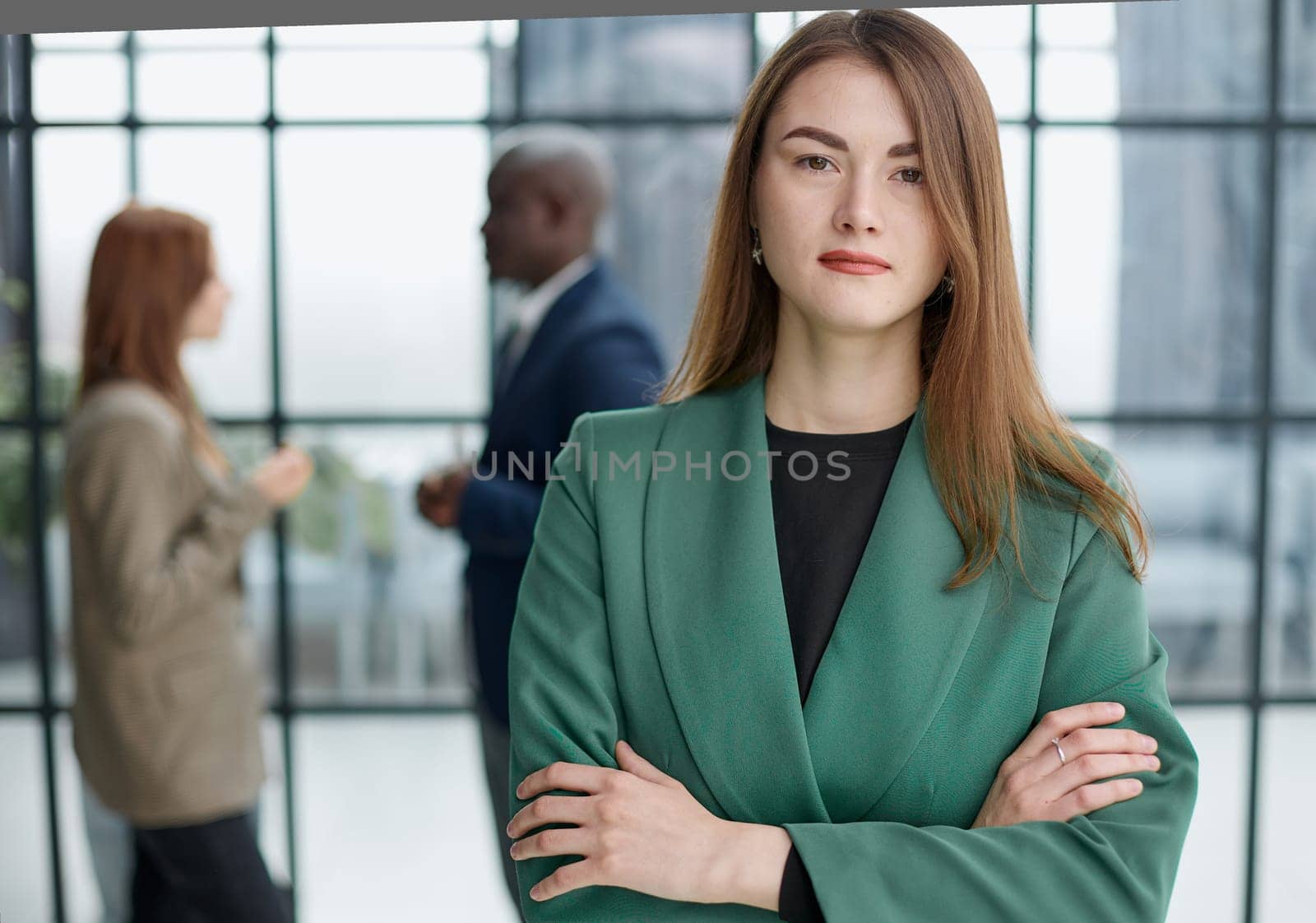 businesswoman standing with her arms crossed in an office while her colleagues have a meeting in the background.