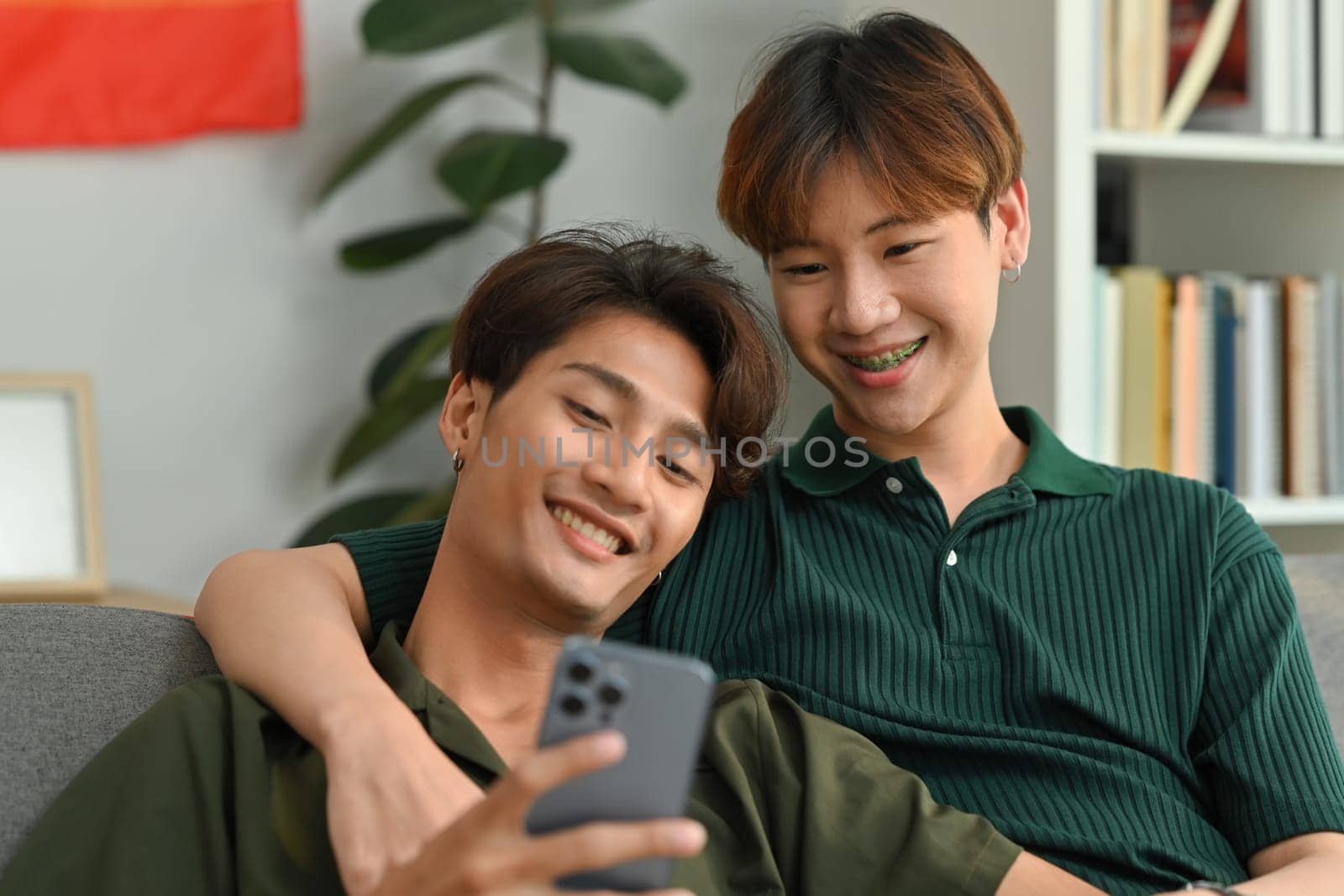 Cheerful two men couple using smartphone, relaxing on couch at home. LGBT, love and lifestyle relationship concept by prathanchorruangsak