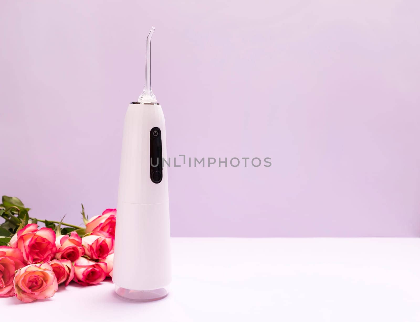 Dental discount, sale, holiday concept. Oral teeth irrigator, roses on purple background. Water tooth cleaner, white portable rechargeable cordless water dental flosser.Home dental care device. Vertical