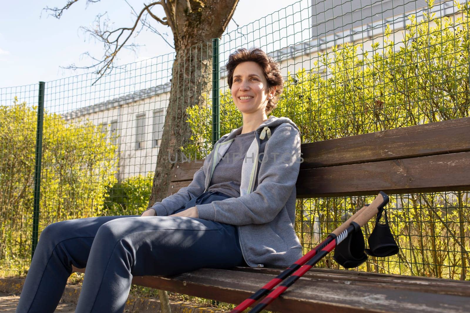 Smiling with Teeth Woman Sits on Bench Holding Poles After Nordic Walking. Beautiful Happy Caucasian Woman Enjoys Sunny Day. Physical Activity With Walking Poles. Outdoor Recreation. Horizontal by netatsi