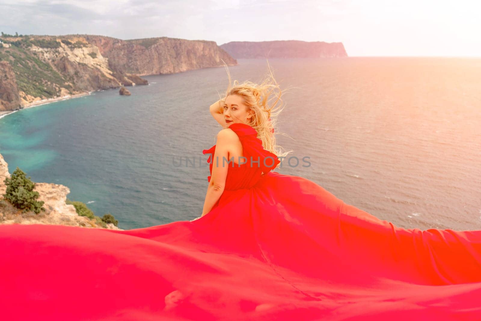 woman red silk dress sits by the ocean with mountains in the background, her dress swaying in the wind. by Matiunina
