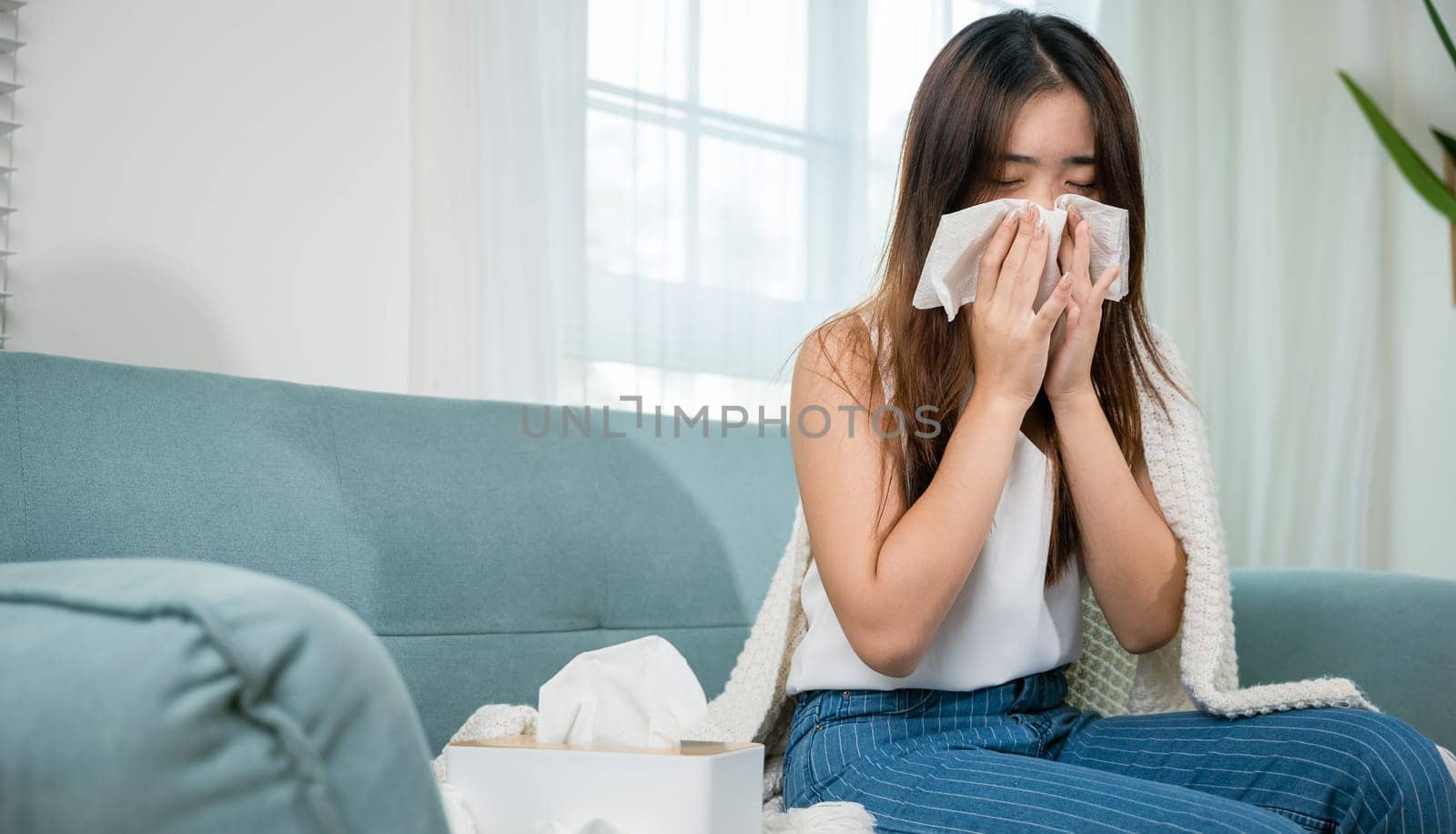 Sick woman sitting under blanket on sofa and sneeze with tissue paper in living room, Asian young female blowing nose sneezing in tissue at home, fever caught cold