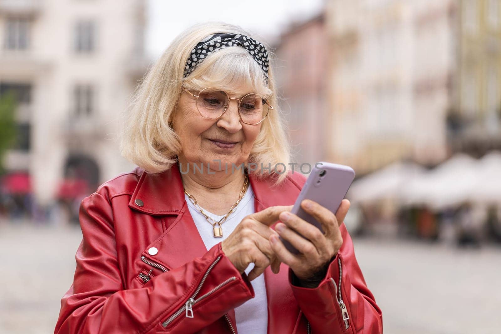Portrait of senior woman using smartphone typing text messages looking for a way on map in mobile navigator app outdoors. Elderly grandmother walking in urban city street background. Town lifestyles