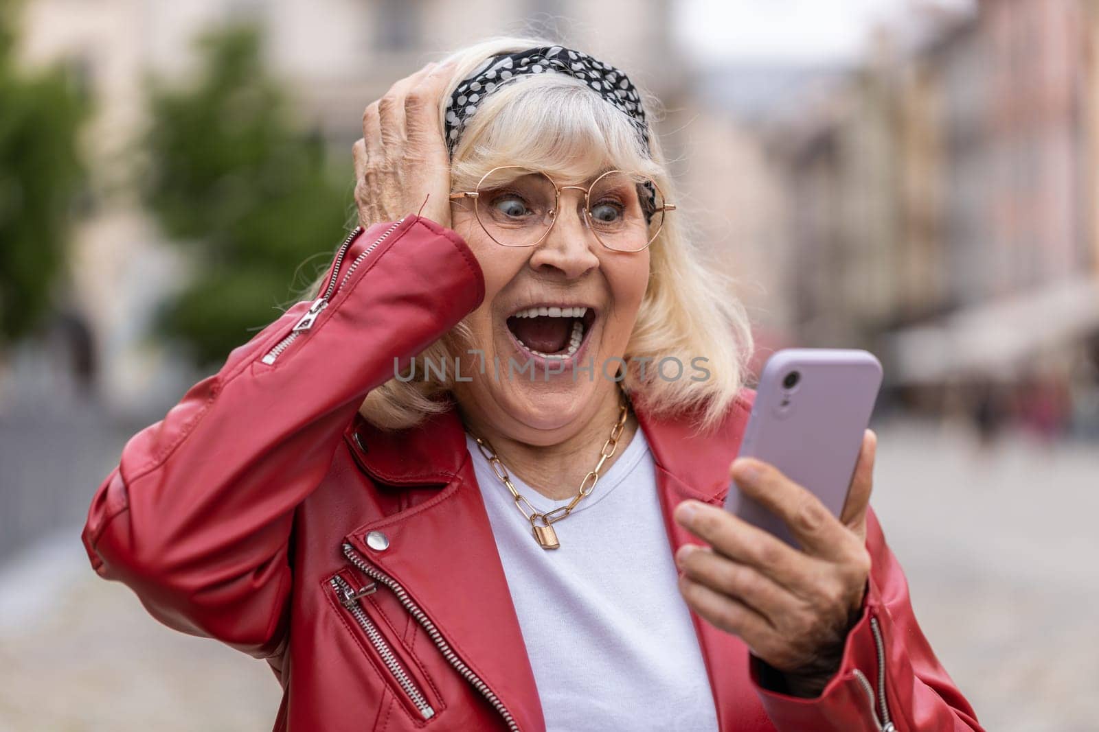 Oh my God Wow. Excited happy joyful winner senior woman use smartphone typing browsing celebrating win good message news. Elderly grandmother with mobile phone walking in urban city street outdoors