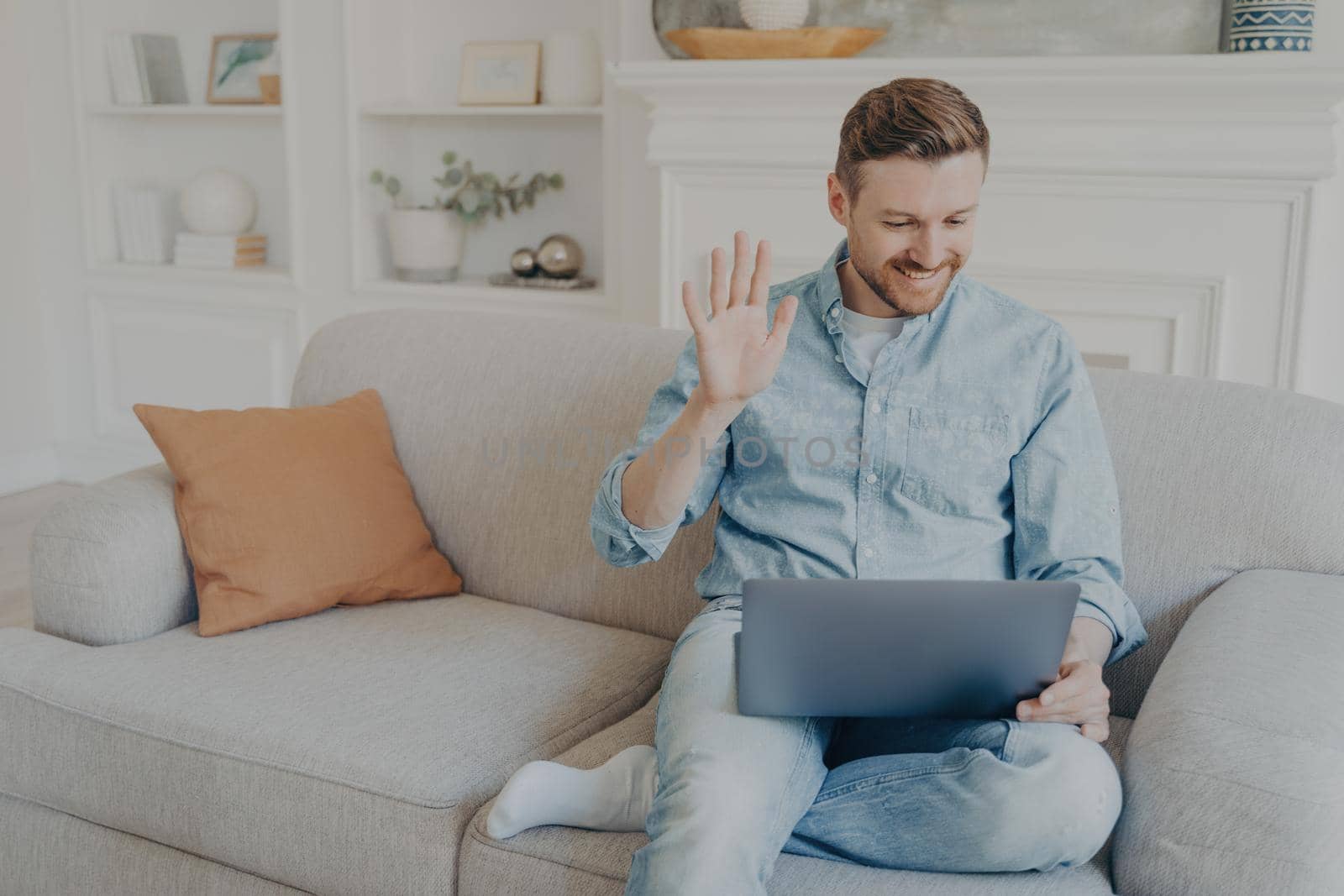 Young handsome casually dressed man waving his hand while speaking with family online using notebook, happy to have conversation with relatives, sitting on couch with leg crossed