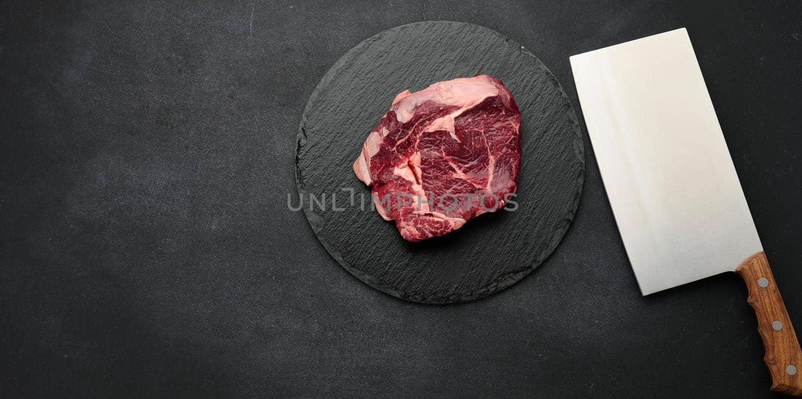 round raw piece of beef meat on a black board. Ribeye for barbecue by ndanko