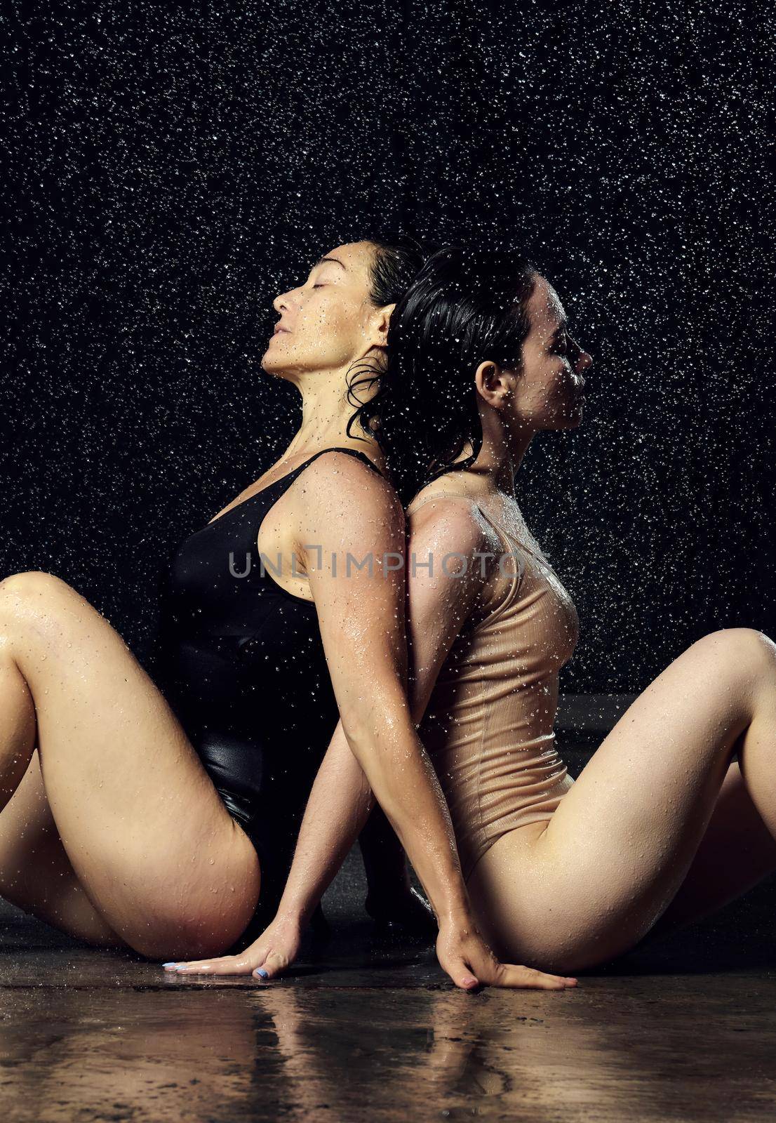 two beautiful women of Caucasian appearance with black hair sit back to back in drops of water on a black background. Women with sad faces by ndanko