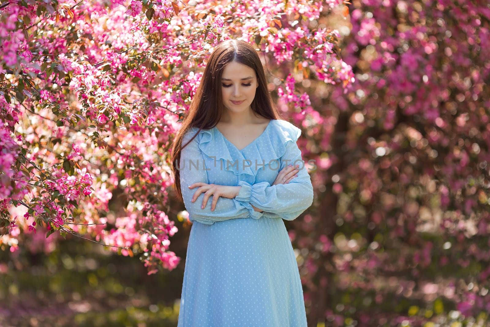 happy brunette girl in light blue dress, with long hair is standing near a pink blooming apple trees, in the summer in the garden. Copy space