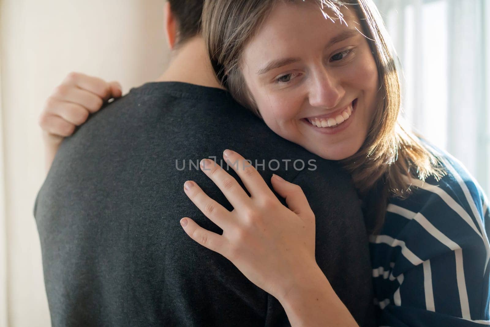 Happy young girl warmly hugs her boyfriend, rejoices at the meeting, stands near the window, smiley face in close-up, woman starts a romantic relationship. Husband and wife feel united, in a harmony.