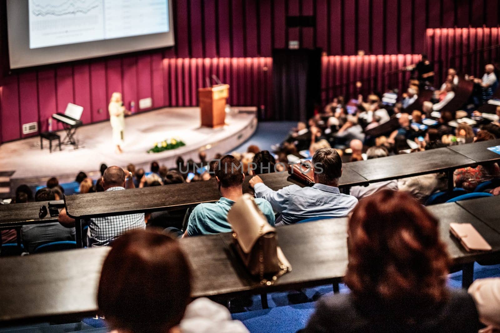 Business and entrepreneurship symposium. Female speaker giving a talk at business meeting. Audience in conference hall. Rear view of unrecognized participant in audience.