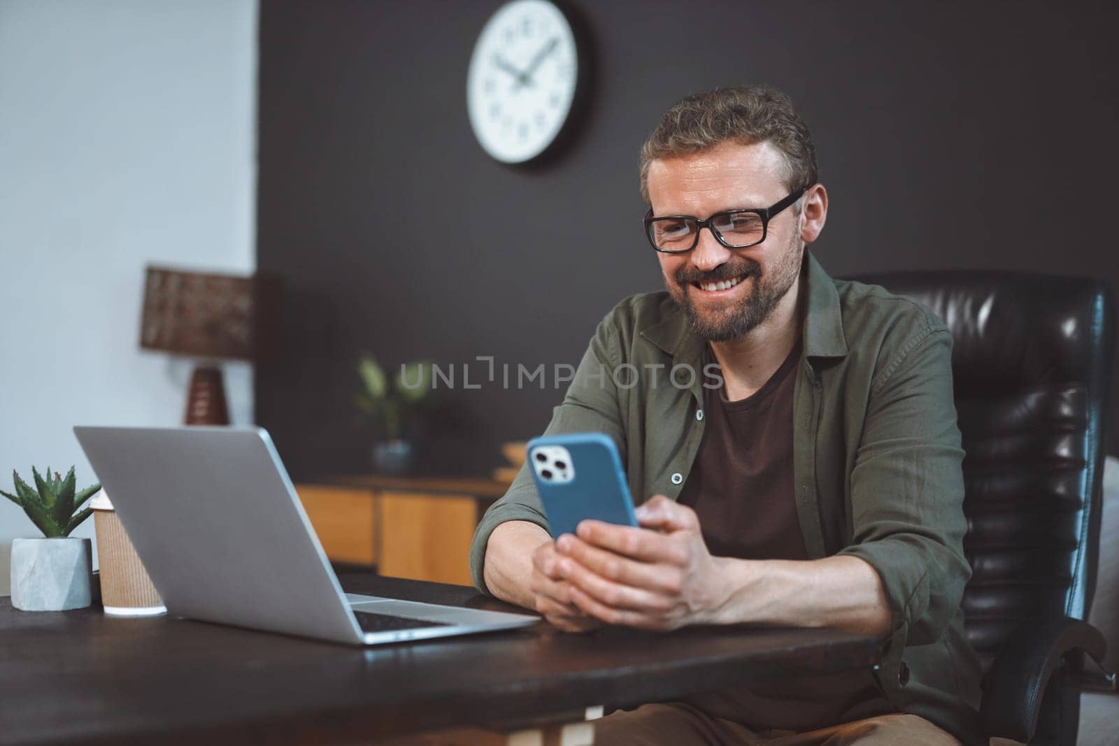Smiling and happy mid-aged man engages in online activities while sitting at home on desk near laptop. Man is depicted multitasking, either sending text message or spending time online through phone. by LipikStockMedia