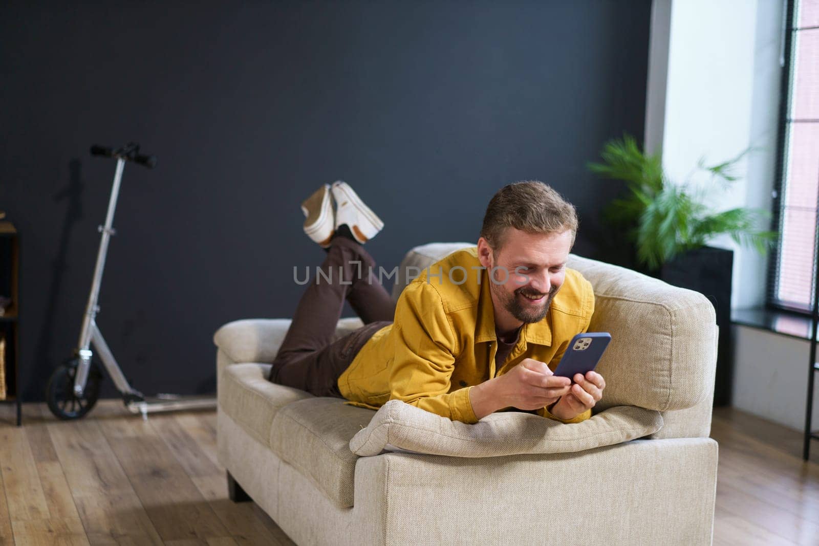 Smiling man enjoying time at home, sitting on sofa with phone. He appears handsome and content engages in texting or messaging on mobile device. High quality photo