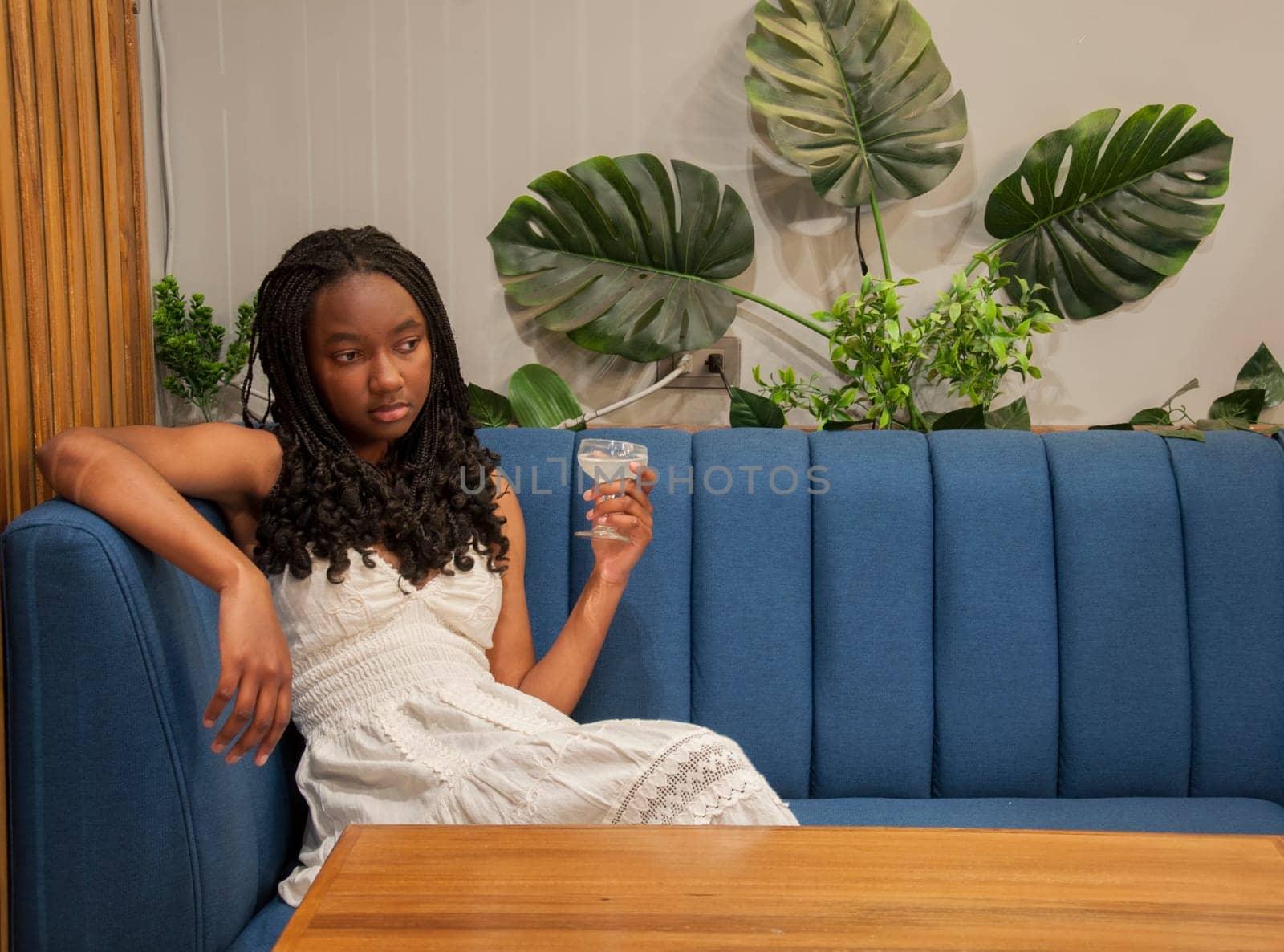 copyspace of young afro man modeling on a blue sofa next to large green leaves of a plant while having a glass of alcochol. High quality photo