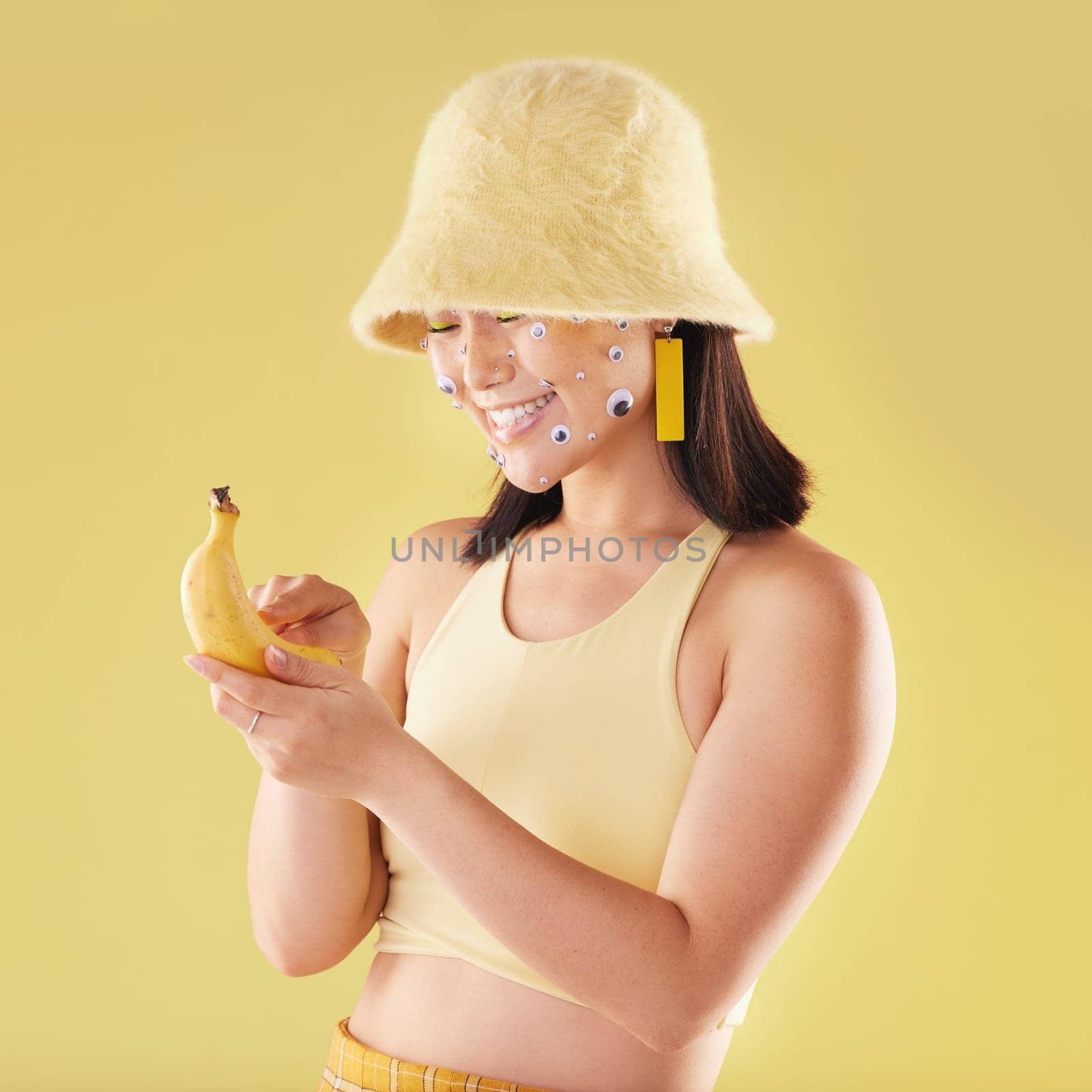 Woman, banana phone texting and studio with smile, communication and eyes sticker art on face. Asian model girl, yellow background and fashion with beauty, aesthetic and cosmetic wellness with fruit by YuriArcurs