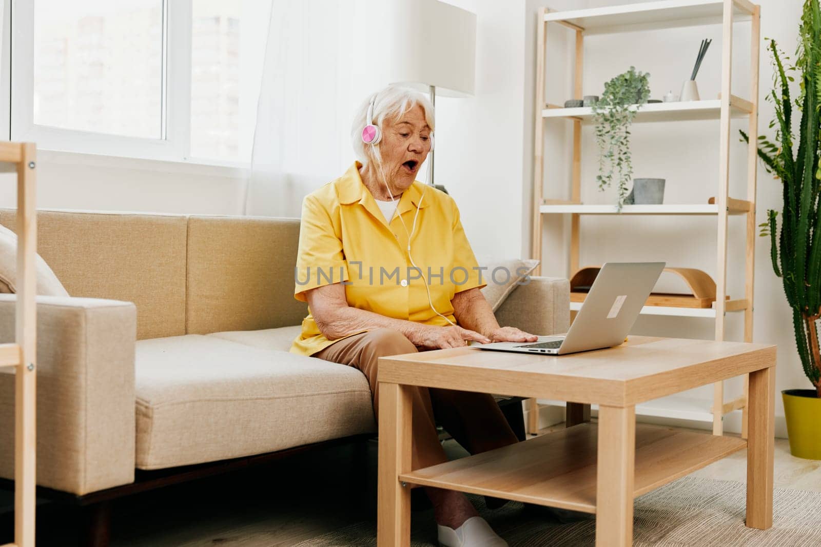 An elderly woman wearing headphones with a laptop sitting on the couch at home and working in a yellow shirt in front of a window, surprise, the lifestyle of a pensioner. by SHOTPRIME
