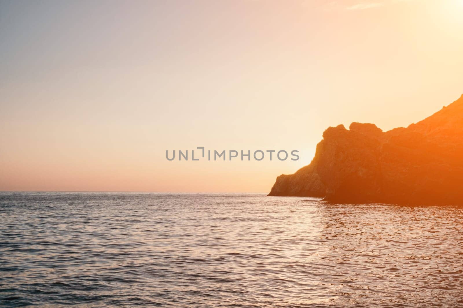 A red burning sunset with the silhouette of a cliff over the sea. Abstract nature summer or spring ocean sea background. Small waves on golden warm water surface with bokeh lights from sun.