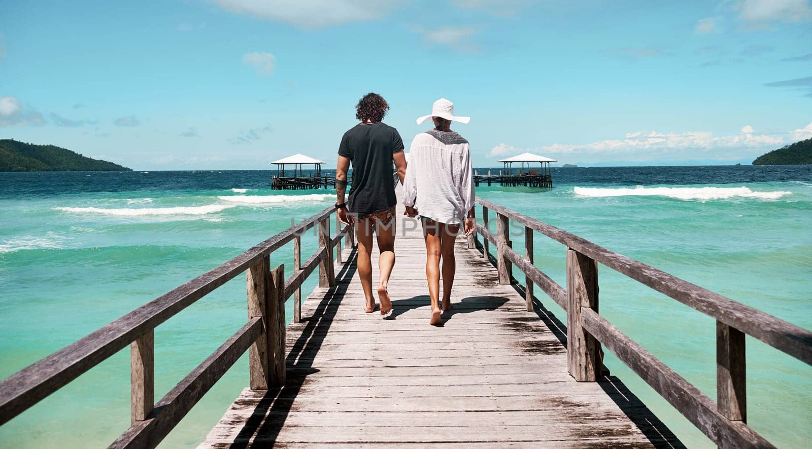 Travel, freedom and rear view of couple at beach walking, holding hands and relax on ocean jetty with blue sky background. Summer, holiday and behind man with woman at sea, traveling or Bali romance.