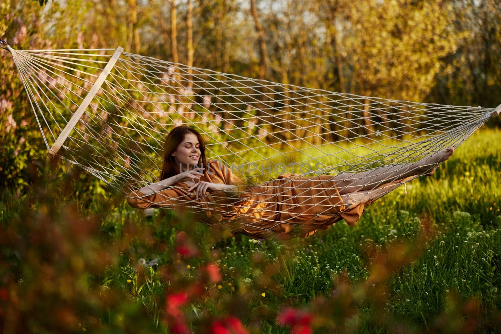 a beautiful woman is resting in nature lying in a mesh hammock in a long orange dress looking at the camera. Horizontal photo on the theme of recreation. High quality photo