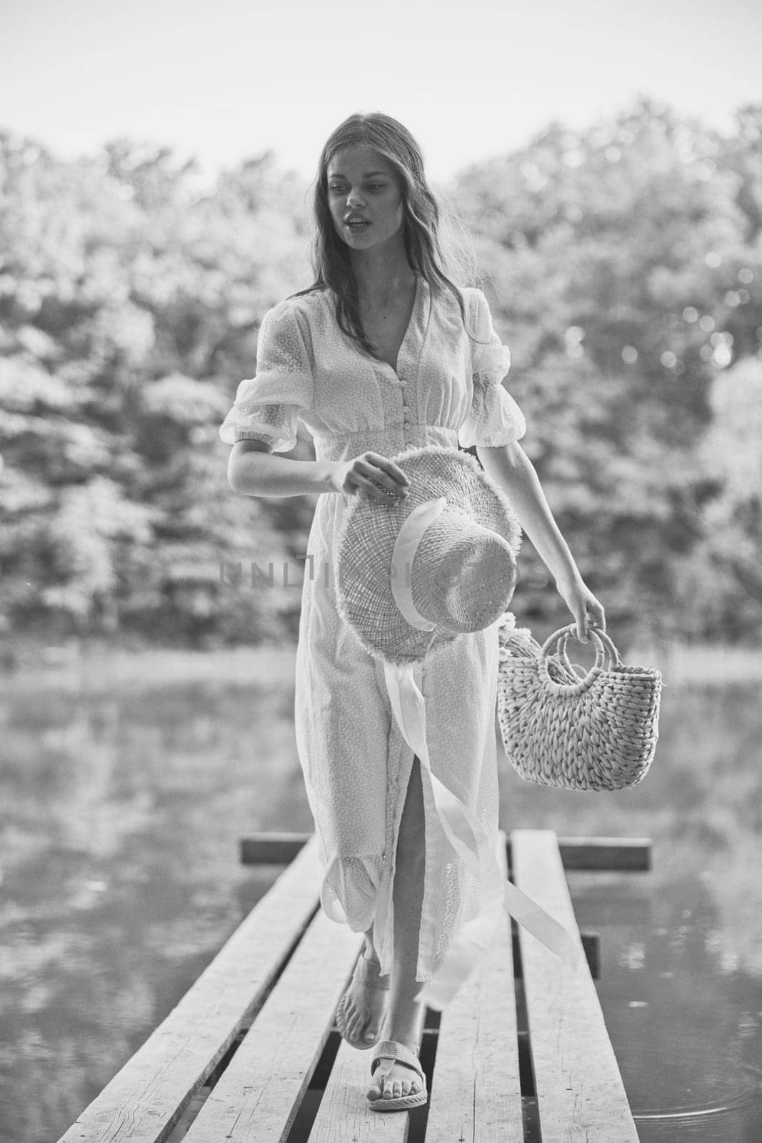 monochrome photo of a woman on the pier of a forest lake with a wicker hat looking at the camera. High quality photo