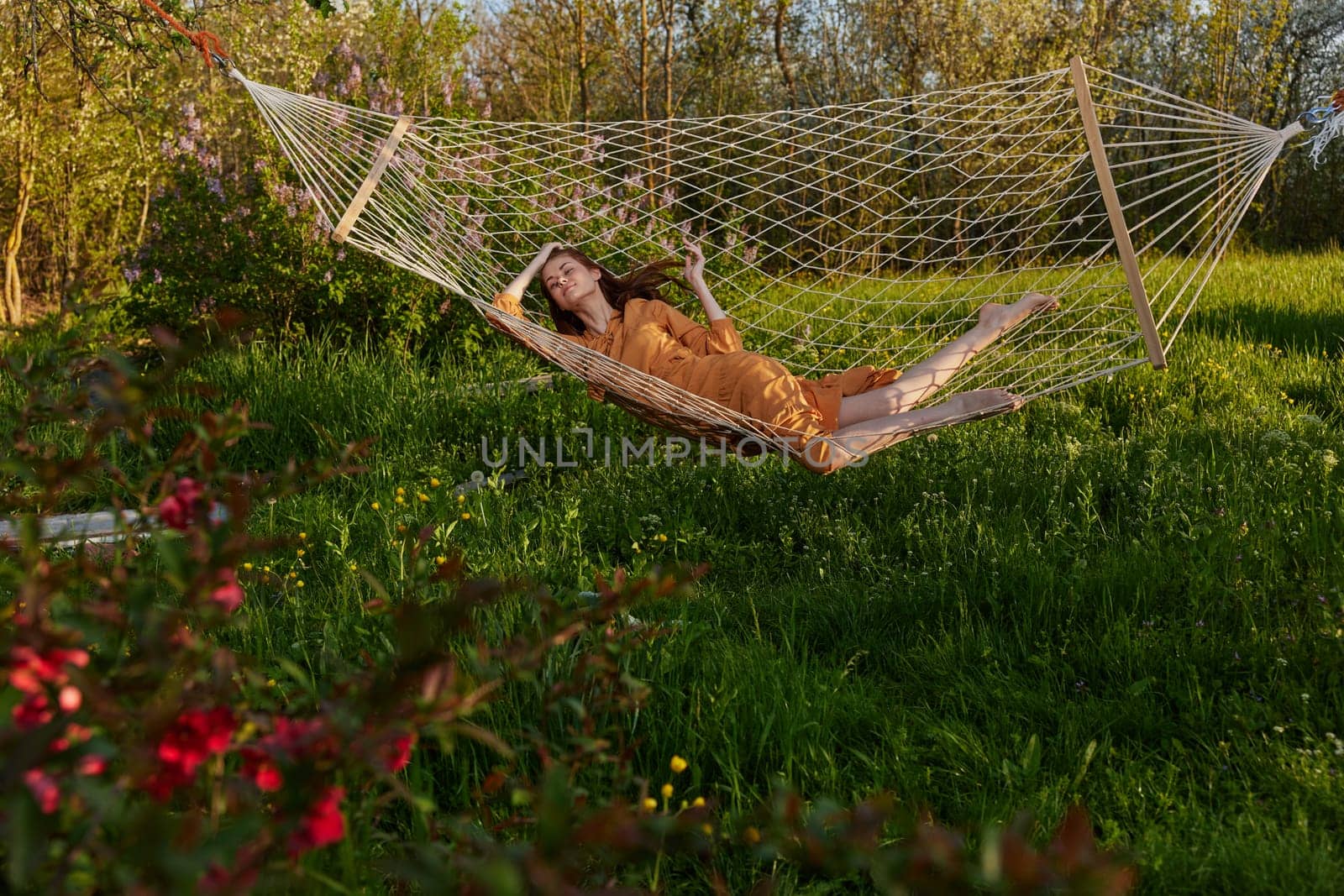 a happy woman in a long orange dress is relaxing in nature lying in a mesh hammock enjoying summer and vacation, looking at the sky by Vichizh