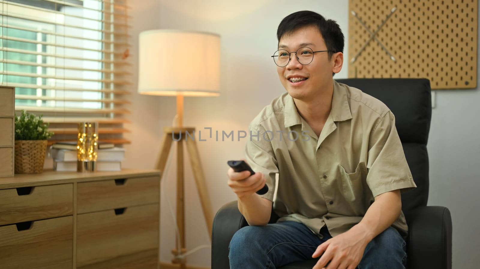 Smiling man in glasses and casual clothes holding remote control, watching TV in cozy living room by prathanchorruangsak