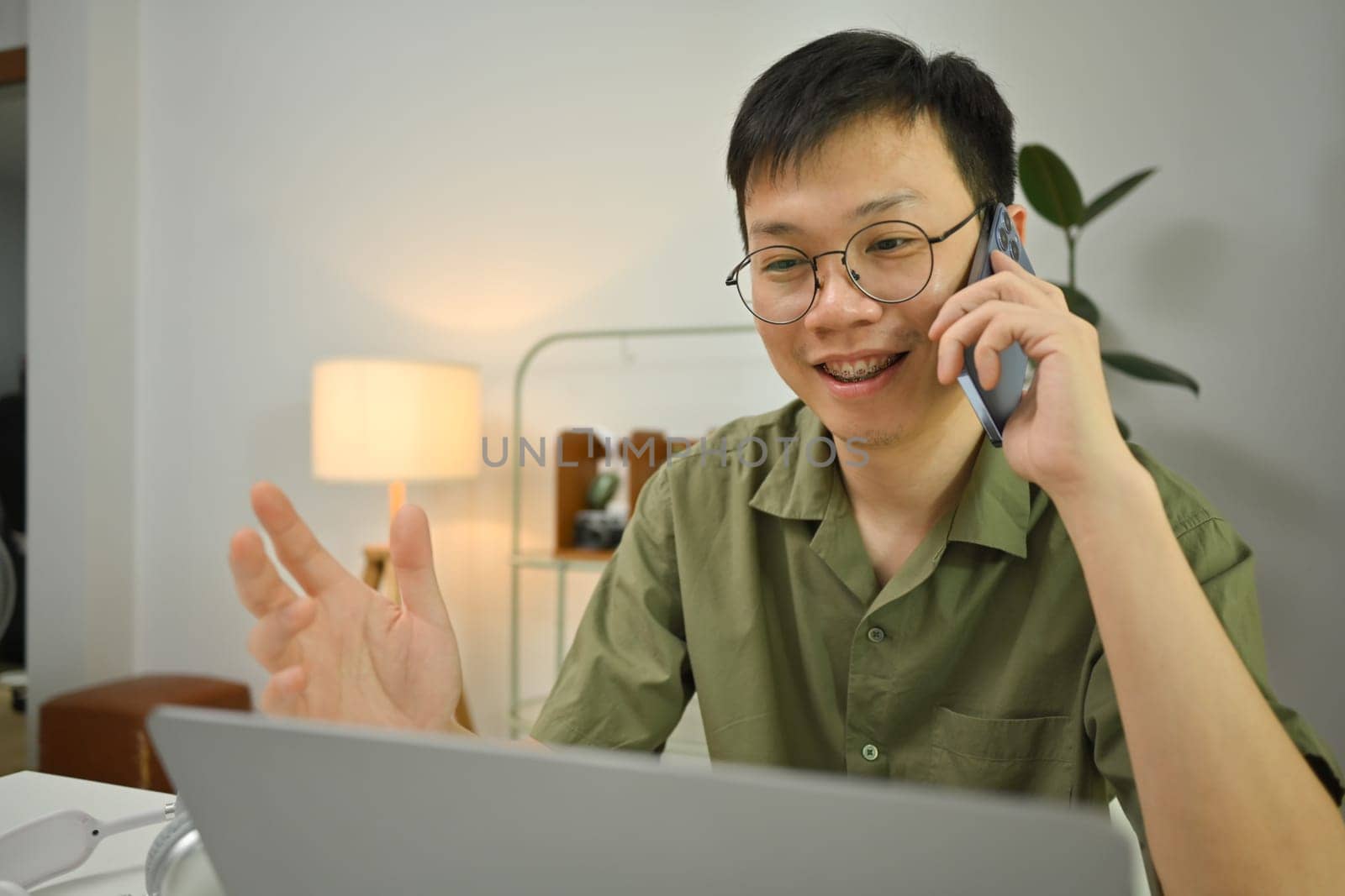 Asian man freelancer in glasses talking on cellphone and looking at laptop screen, sitting at desk in home office.