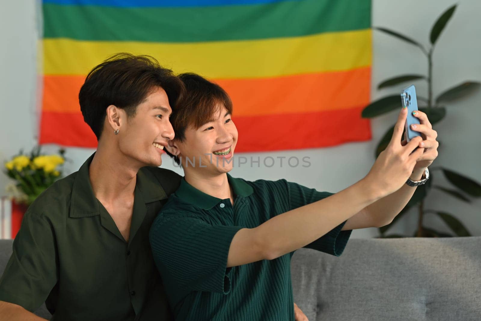 Joyful homosexual couple taking a selfie with smartphone in living room with rainbow flag in background. LGBT and love concept.