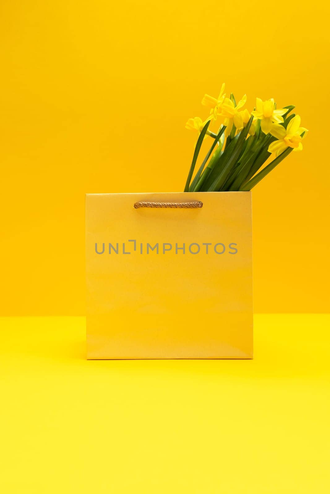 Bouquet of yellow narcissus in golden package, bag on yellow orange background. Spring sale, discount. International Women's Day celebration, Copy space for text. Monochrome minimalism. Vertical