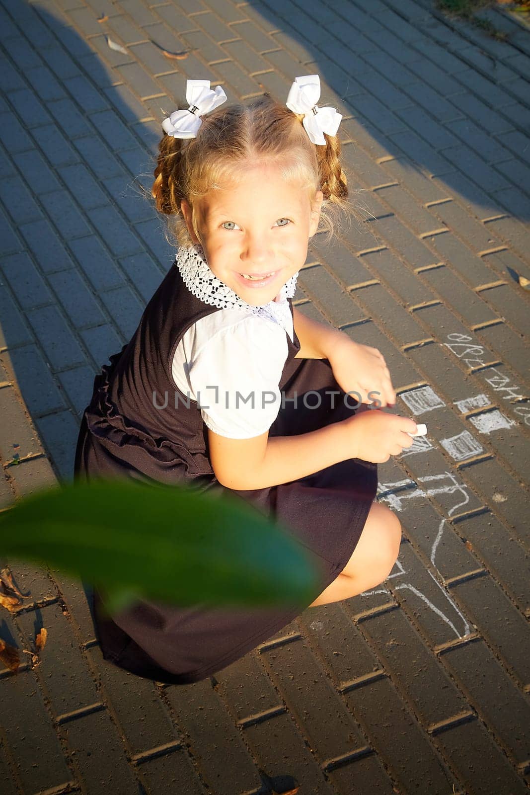 Little girl of elementary school student in modern school uniform drawing with chalk on asphalt outdoors. Female child schoolgirl going to school. Back to school in september 1 in Russia by keleny