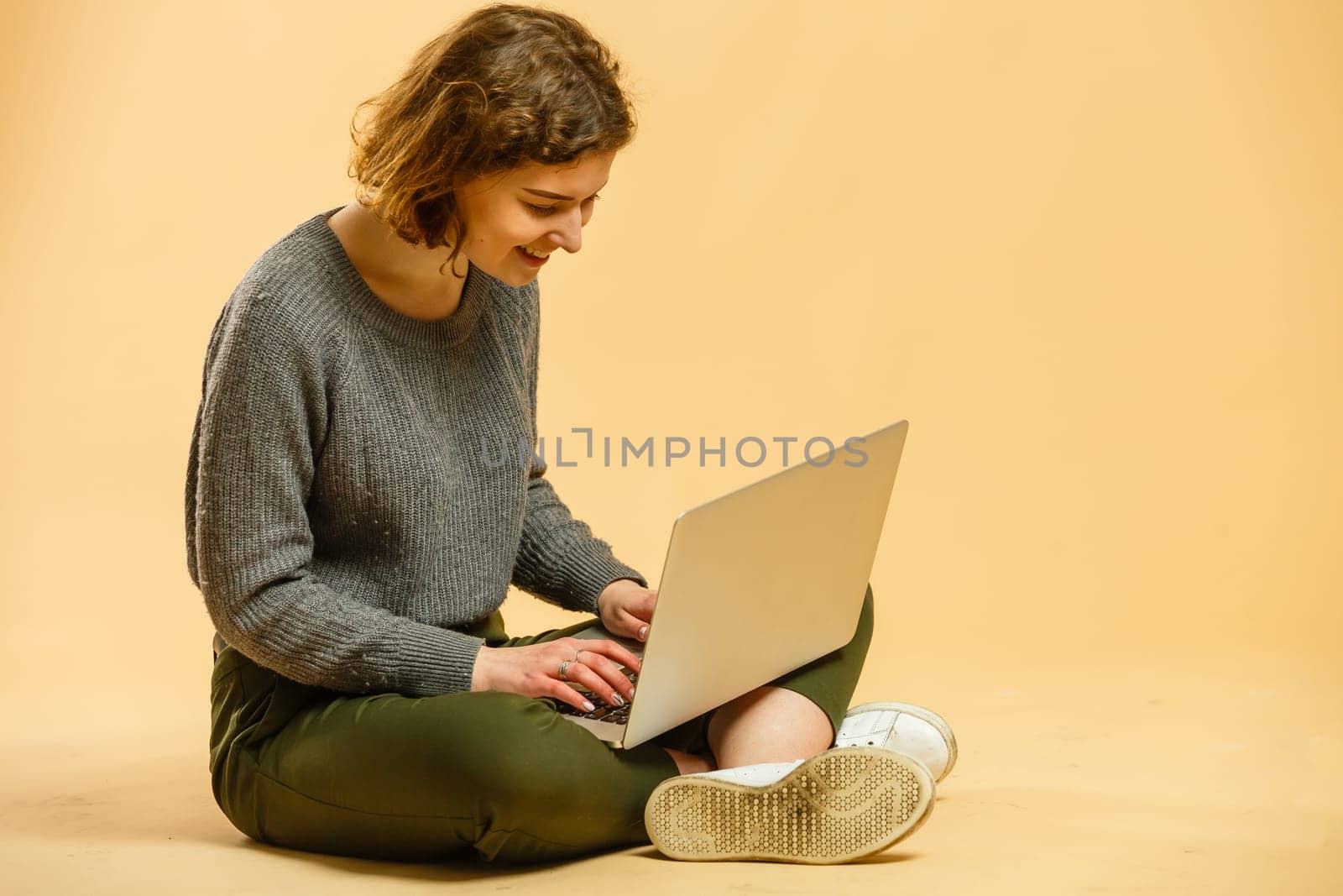 Happy young woman sitting on the floor with crossed legs and using laptop on beige background