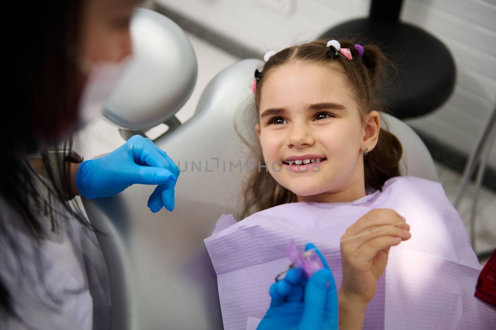 Multi ethnic little kid girl in dentist's chair, smiles broadly to her doctor dentist hygienist during regular dental check-up at pediatric at dental clinic. Pediatric dentistry. Child teeth therapy