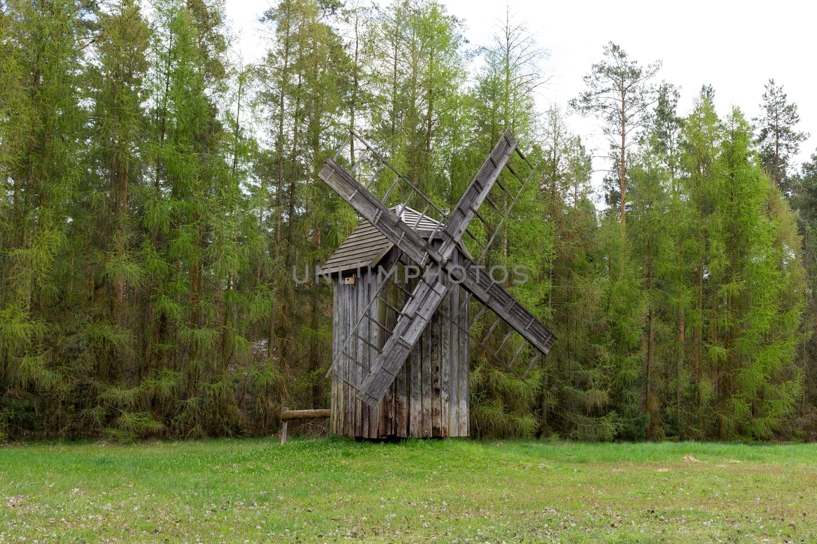 Old Wooden Mill In Meadow, Open Air. Blades Of Old Wooden Windmill In Rural Eastern Europe Area, Countryside. Green Forest On Background. Horizontal Landscape Plane High Quality Photo.