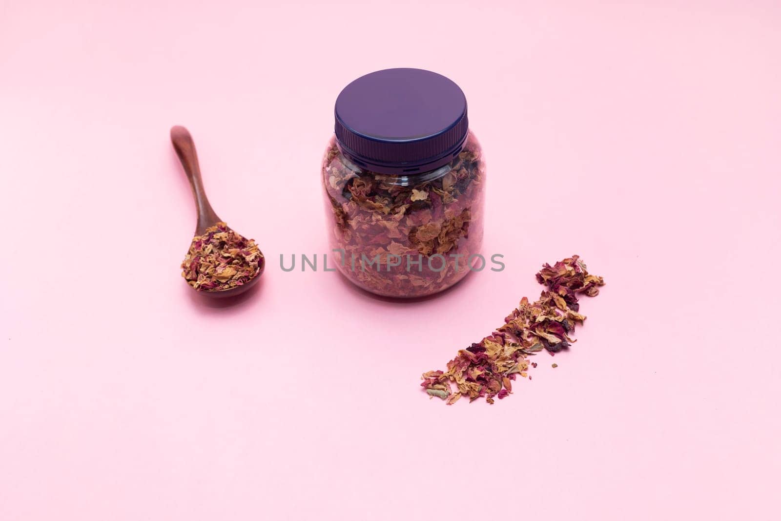 Jar, container with dry roses petals, leaves. Wooden spoon filled with dried roses on pink background. Aromatic herbal beverage made from fragrant buds of flowers. Top view,flatly horizontal.Copyspace