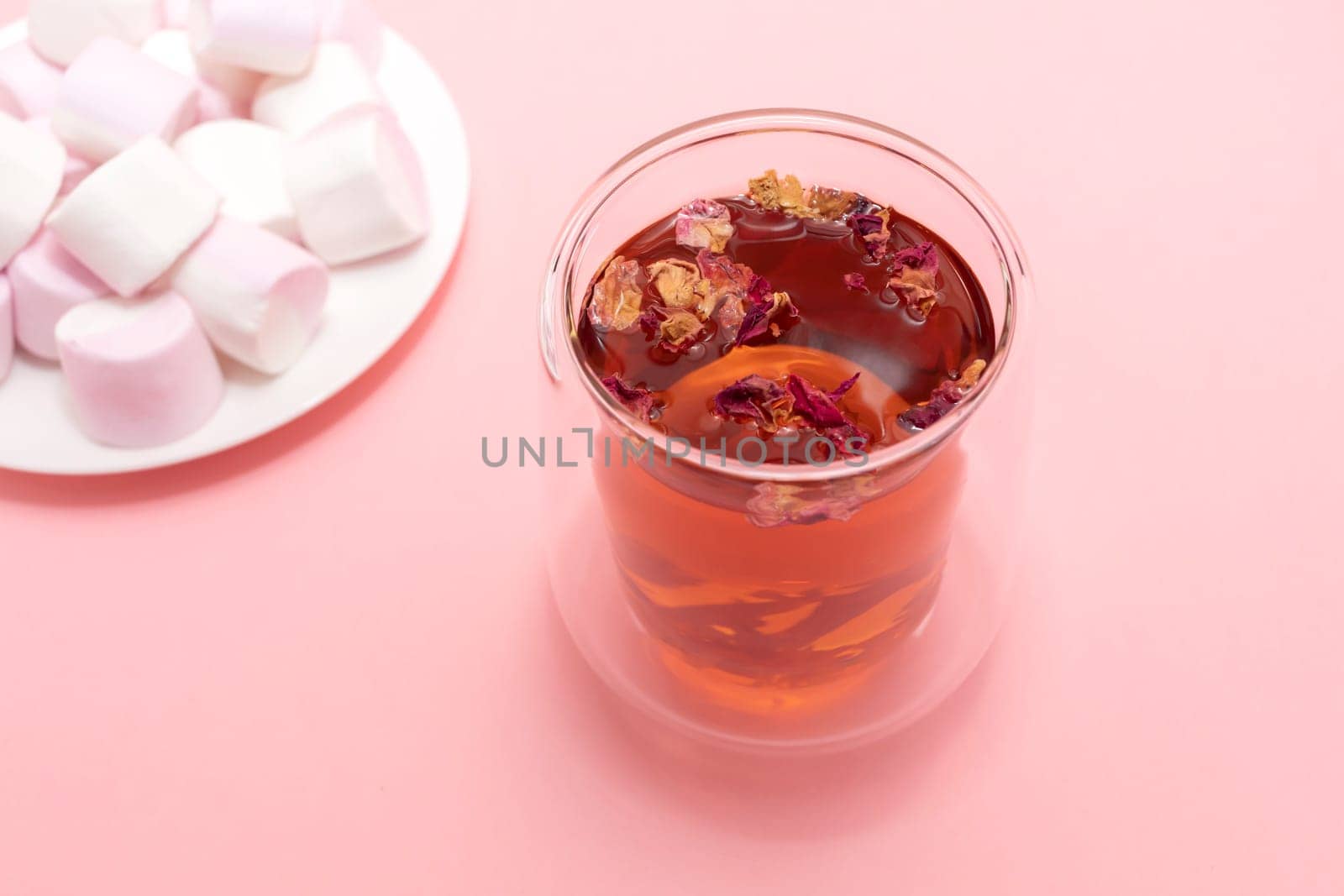 Healthy herbal rose petals, buds tea in double walled glass mug, cup. Pink white marshmallow plate. Pink background. Desiccated colorful flower petals herbal drink, beverage. Horizontal, top view.