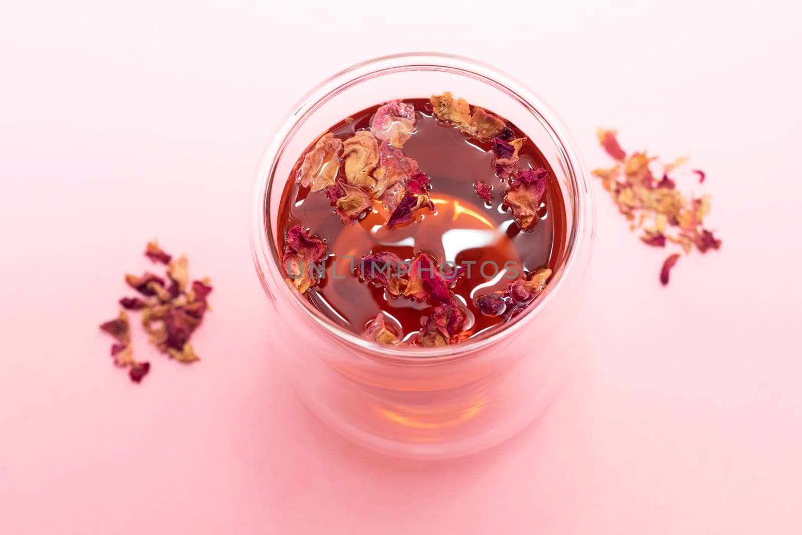 Healthy herbal rose petals, buds hot tea in double walled glass cup on pink background. Desiccated colorful flower petals herbal drink, beverage. Horizontal, top view, closeup.