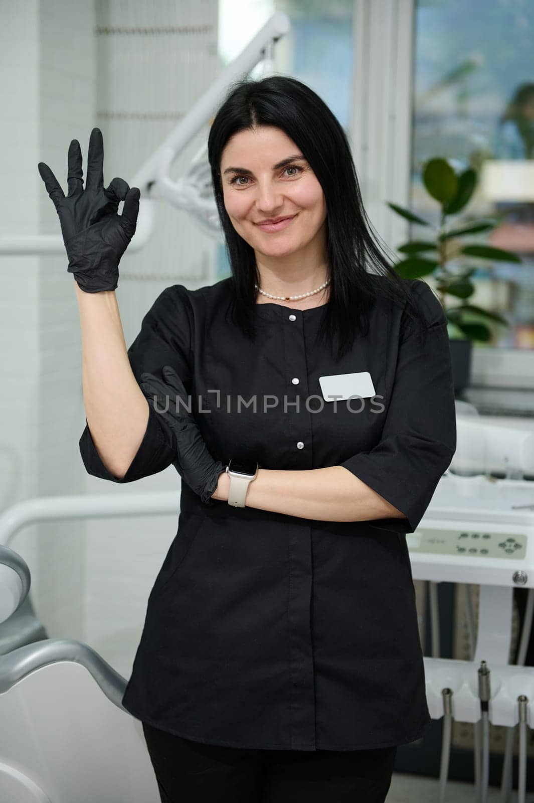 Professional portrait of young pretty woman dentist doctor in stylish black uniform, standing near the dental chair in the office, gesturing with Ok hand sign, smiling, confidently looking at camera.