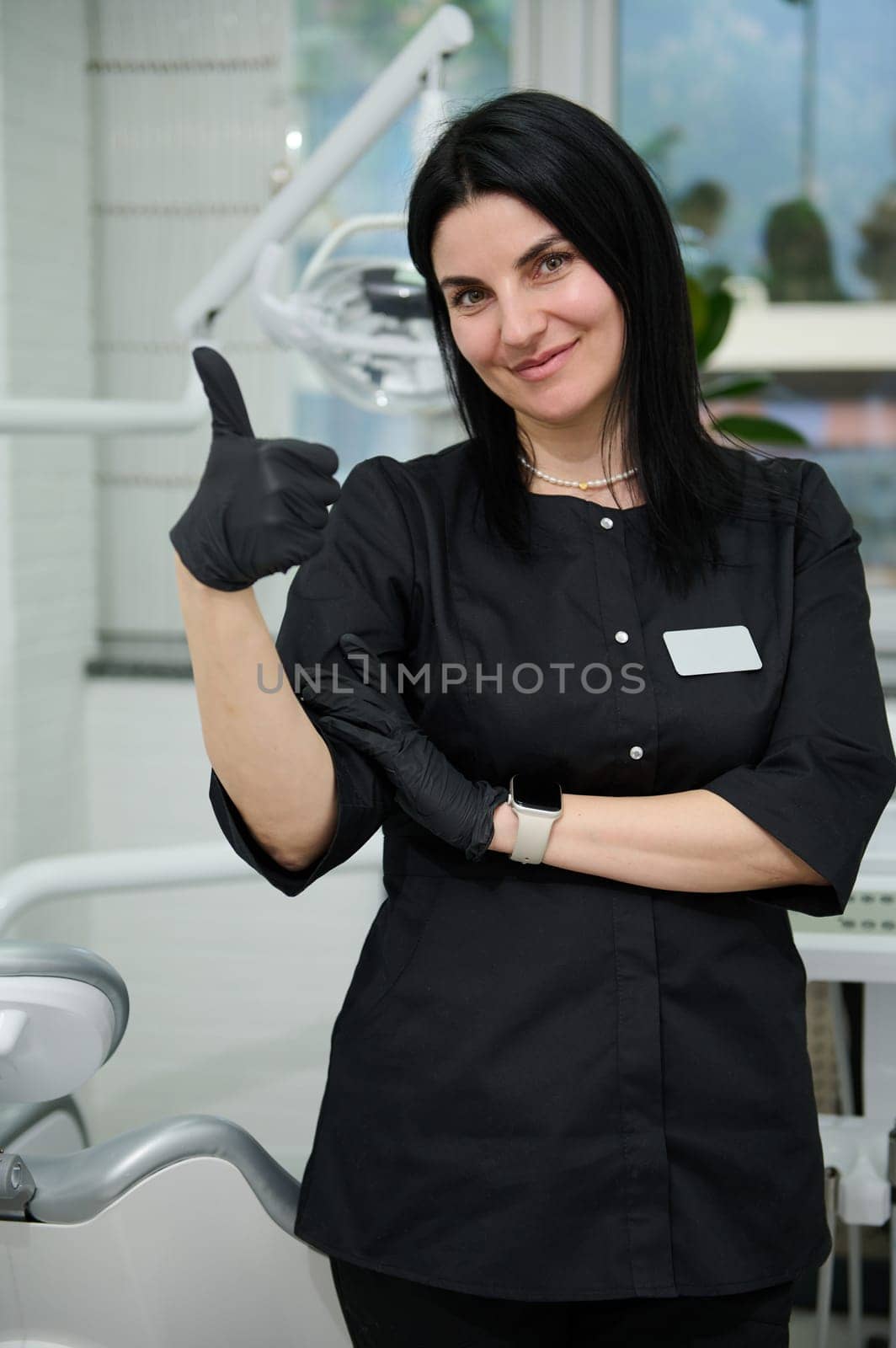 Portrait of charming Caucasian middle aged woman dentist hygienist, dressed in black medical uniform, smiling cutely looking at camera, showing thumb up, standing in her office at newest dental clinic
