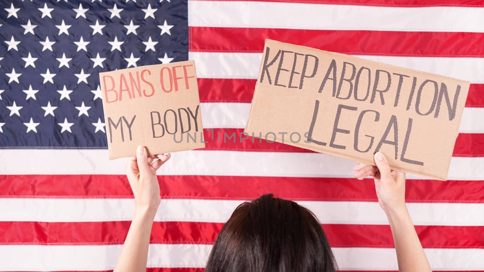 Young woman protester holds cardboard with Keep Abortion Legal and Bans Of My Body signs against USA flag on background. Girl protesting against anti-abortion laws. Womens rights freedom