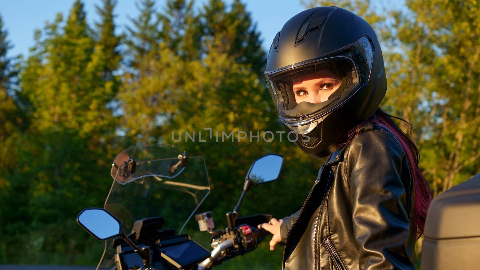 Young biker girl riding touring motorbike on forest road. Beautiful woman with red hair in leather jacket sitting on motorcycle in the park